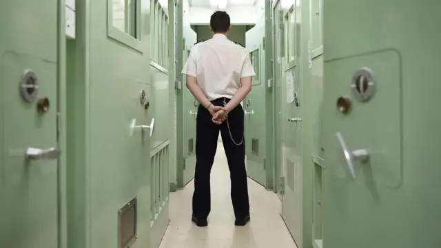 A prison guard stands in a narrow prison corridor with green doors