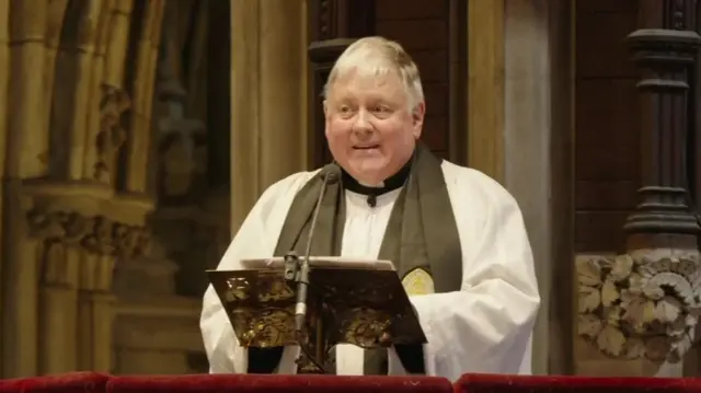 A priest in white robes and black scarf around his neck. He has short white hair and stands on a plinth reading from a bible.