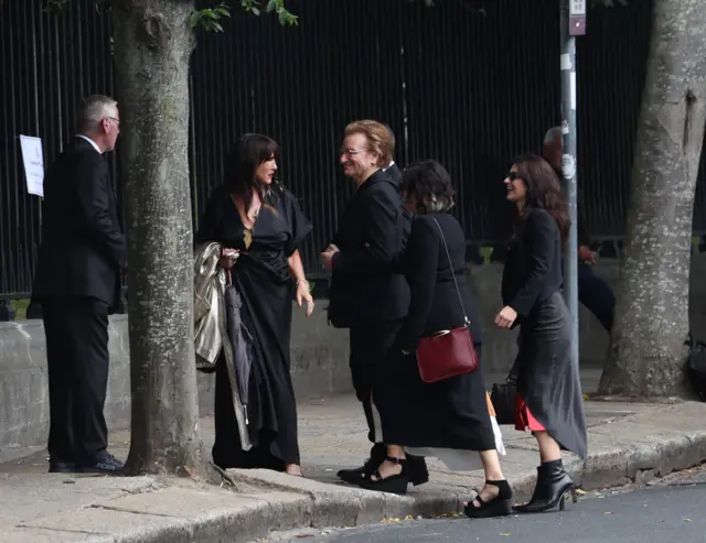 Bono, a man with brown hair and glasses, arrived outside the church wearing a black suit. He was accompanied by women who were also wearing black.