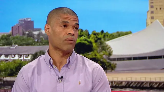 Calvin Buckley, a man with close cropped hair, wears a shirt sitting on the BBC Breakfast set