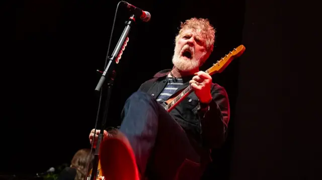A man with short grey hair and a short grey beard hold a guitar with both hands. He wears a black jacket with a navy and white striped t-shirt. He sings into a black and silver microphone