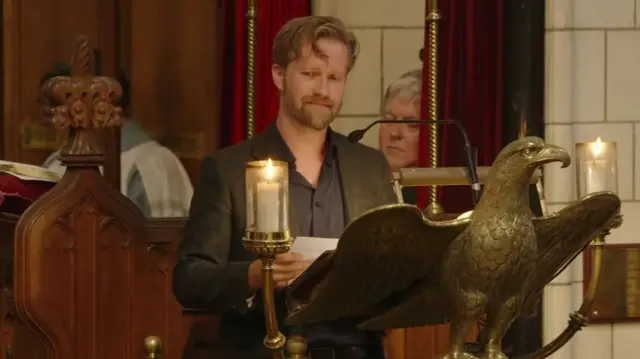 A man with short red hair stands behind a golden eagle. He wears grey suit jacket and navy suit.