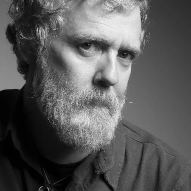 A black and white portrait photo of Glen Hansard, a singer with short curly hair and a greying beard. He is wearing a dark shirt and a necklace