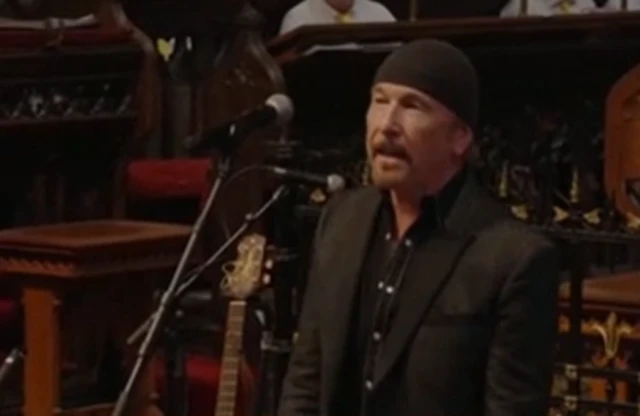 The Edge, a man with a black beanie cap and dark suit, sings into a microphone in church