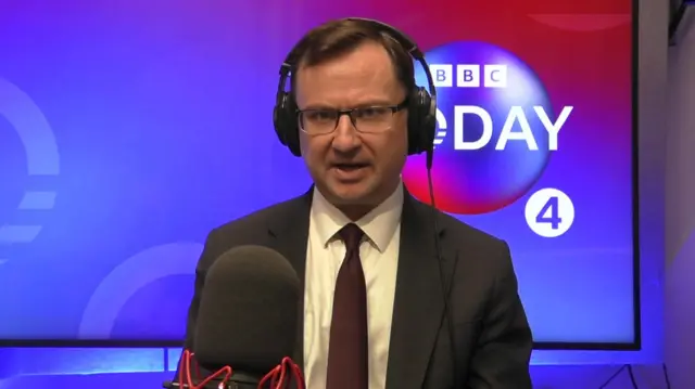 Alex Norris sits in a dark suit and dark red tie while wearing headphones and sitting behind a microphone. The backdrop for Radio 4's Today programme is behind him.