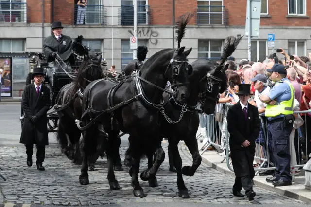 Four black horses pull a carriage containing the coffin of Glen Hansard as a crowd lines a street.
