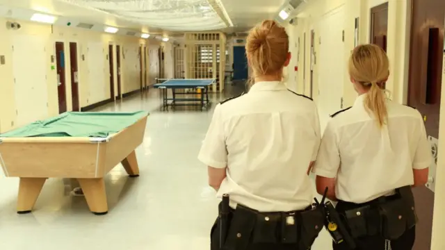 Two women working in a prison, walking down the corridor, with white tops