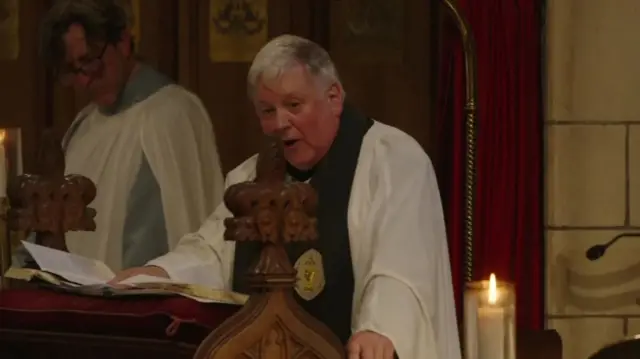 A priest in a white robe stands at a wooden alter. A priest in white robe stands to his right.