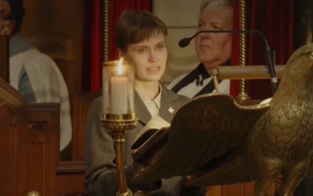 Maire Saaritsa, a woman with short, dark hair, speaks into a microphone during the funeral