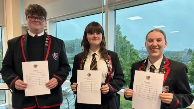 Three school pupils, a boy and two girls, stand side by side in school uniforms holding exam results