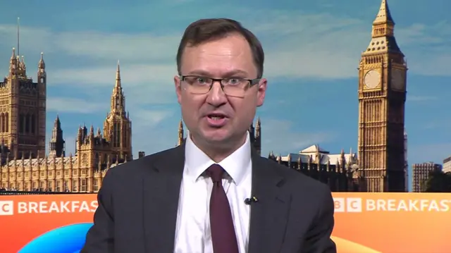 Alex Norris sits in a dark suit and dark red tie in front of the backdrop for BBC Breakfast