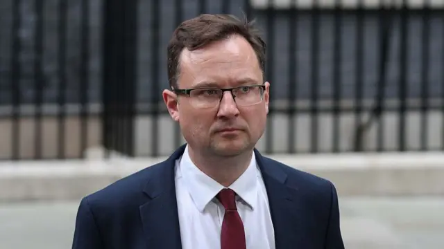 Alex Norris in a navy suit and red tie. He appears to be walking on Downing Street.
