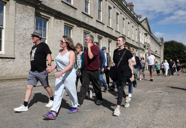 A dozen fans walk in front of a grey stone building.