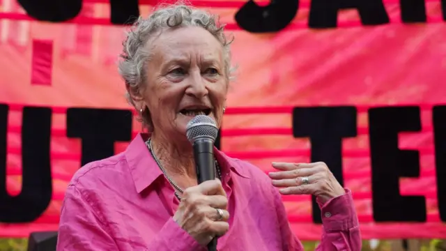 A woman with short grey hair speaks into a microphone in front of a pink banner. She is wearing a pink shirt