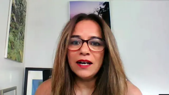 Pia Sinha sits in a room with white walls while wearing glasses. There are two paintings hung on the wall behind her, one green and another purple.