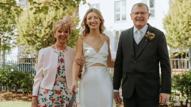 Claire and her parents on her wedding day, smiling in a park