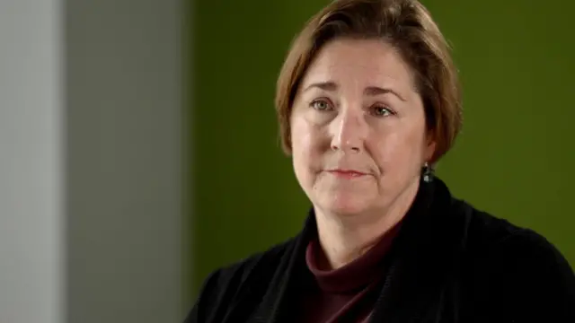 Nicola Jacobs, who has short brown hair, in a close-up shot. She is wearing a dark jacket over a red top, with a green wall in the background
