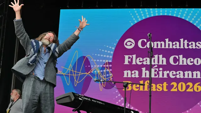 A man is standing onstage with his arms up. He has long hair and a beard and wears glasses. He's wearing a grey suit.