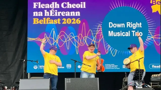 Three men with yellow tee shirts holding irish traditional instruments are onstage perfoming. Two have their hands in the air, all are wearing purple hats and blue jeans.