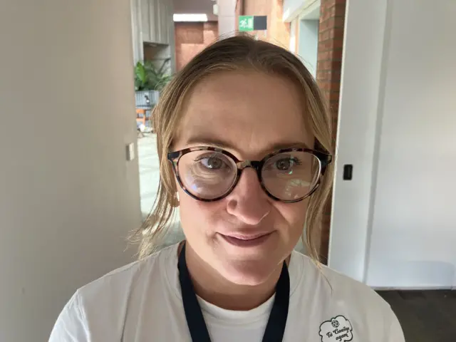 A woman with blonde hair and round black glasses. She wears a white tee shirt with a black lanyard around her neck