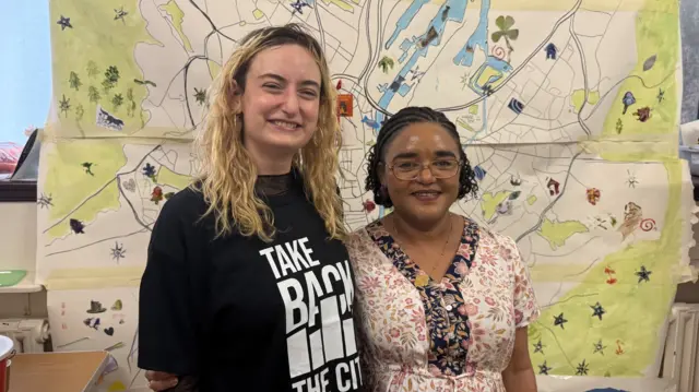 Two women smile at the camera, the women to the left is wearing a black tee shirt and white writing. She has curly blonde hair. The women on the right is shorter and is wearing glasses and flowery pink and white top and has dark braided hair.