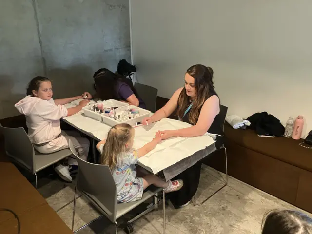 The two children are getting their nails polished by (sitting opposite) two adults sitting at a table. In the middle of the table there is a box filled with colourful polish and makeup