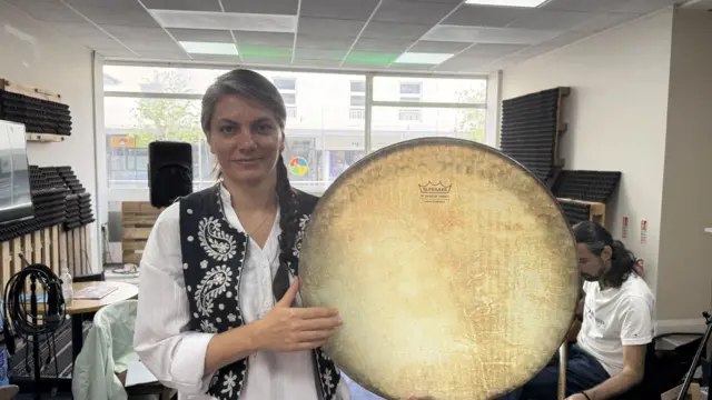 A woman wearing a white shirt, and a black waistcoat with white artwork on it. She is holding a large brown drum in both hands.