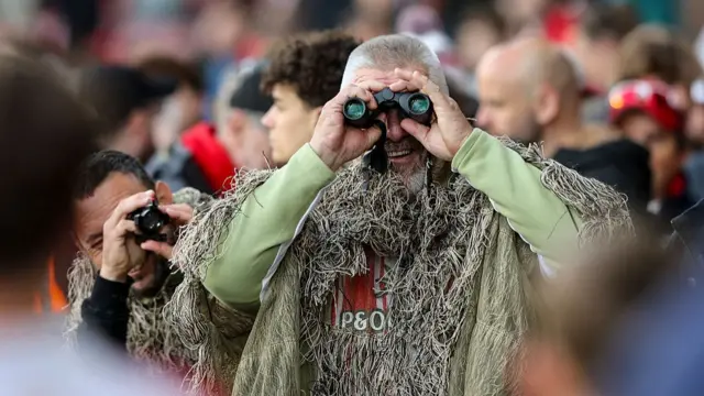 A man in camouflage clothing holding binoculars and wearing a Southampton Football Club t-shirt in a crowd of people