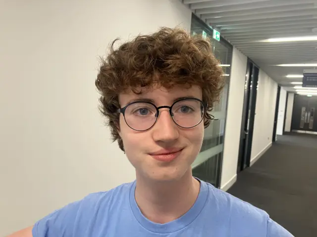 A teenager with red curly hair, round black glasses and a blue tee shirt smiles at the camera.