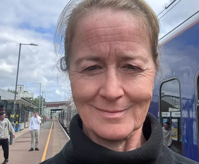 Sarah Nelson, looking into the camera and standing on a rail platform wearing a black high neck sweater