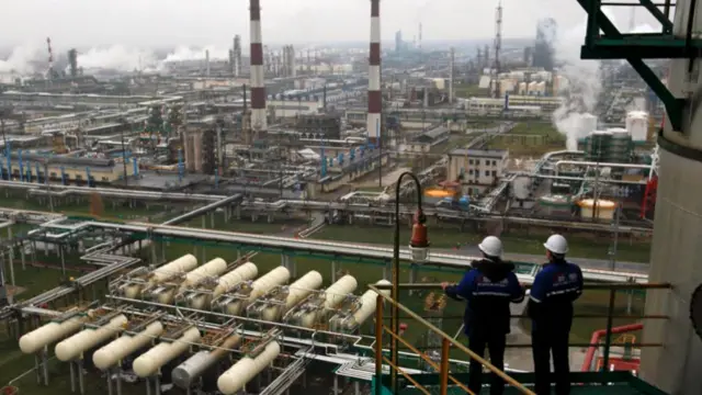 Two men with hard hats on stand on a balcony and look out, surveying a huge industrial complex which has tanks and chimneys with smoke rising