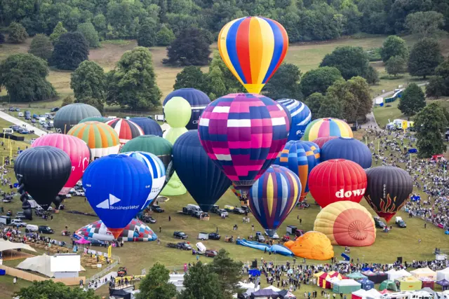 A number of brightly-coloured hot air balloons inflate and lift off from parkland, surrounded by crowds.