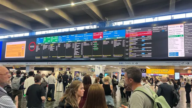 Wide shot of crowd observing the departure board at Euston Station showing a red warning about disruptions
