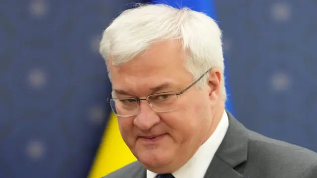 Andriy Sybiga wears a grey suit with a Ukrainian flag visible in a blurred background