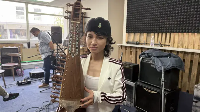 A woman wearing a black hat with a white jacket with three purple stripes down the side. She is holding a Rubab, a traditional guitar from Afghanistan.