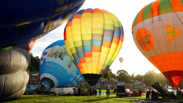 Balloons are inflated in preparation for the mass ascent. One balloon can be seeing flying in the distance. Sunlight is filtering through one multi-coloured balloon.