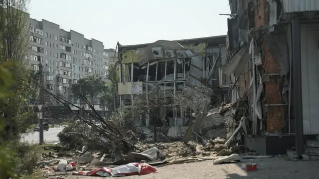 A street showing a ruined multi story building with rubbish all over the street and trees bent over