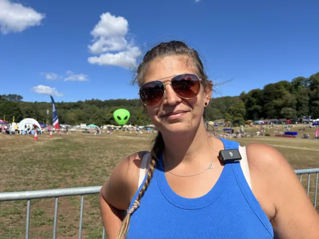 Amy smiles at the camera. She wears a bright blue vest top, large aviator sunglasses and her hair in a long braid.
