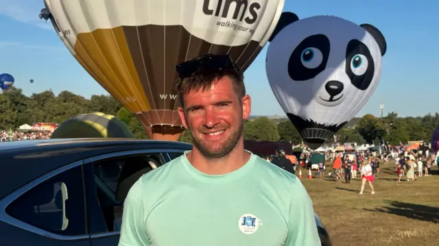 A man stood in a field while hot air balloons take flight behind him. He has short brown hair and facial hair. He is looking at the camera, squinting slightly, but smiling.