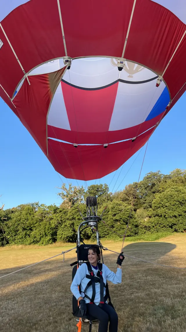 Jo sits in a seat hanging from a hot air balloon. She has dark brown hair and wears a light jumper and leggings, and is smiling while looking away from the camera.