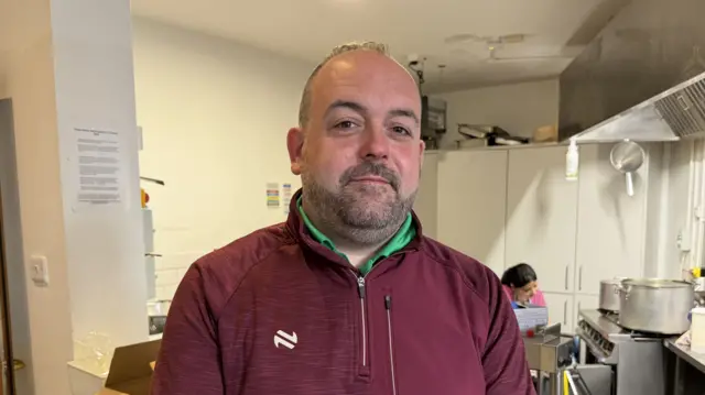 A man with a purple half zip sports top, with a green collared shirt showing underneath. He is bald and has a dark beard. In the background you can see a kitchen with a silver stove, pots and pans .