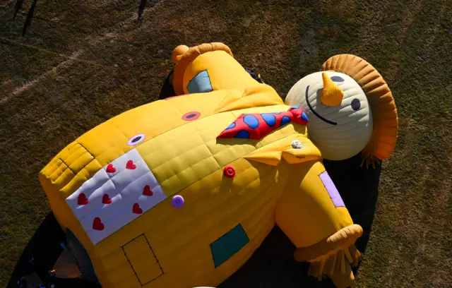 A large yellow hot air balloon shaped like a scarecrow is lying down as it is inflated at the Bristol Balloon Fiesta