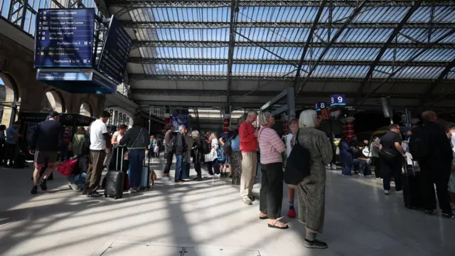 Passengers wait around at Lime Street railway station in Liverpool. Several people with suitcases can be seen looking at their phones while other people look at a departures board