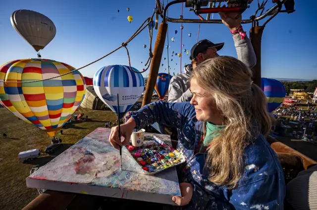 A woman with blonde hair, wearing blue overalls covered in paint, holds a large canvas as she looks out over the view from a balloon basket. There are inflated balloons behind her.