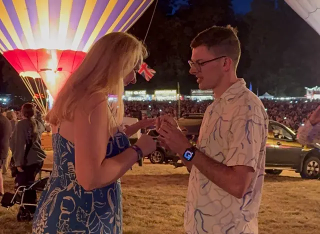 A young man in a pale patterned shirt offers a ring box to a lady with blonde hair. She is wearing a blue and white dress. The man has short dark hair and glasses