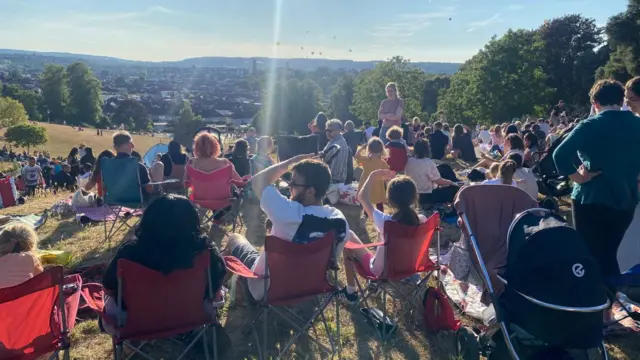 A view taken from behind of crowds in the sunshine watching hot air balloons which are on the horizon