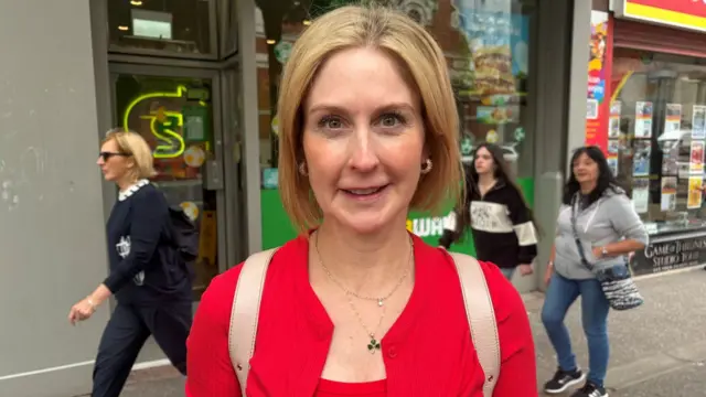 a blonde woman in a red twinset looking at the camera. A high street is visible behind her, with people walking past
