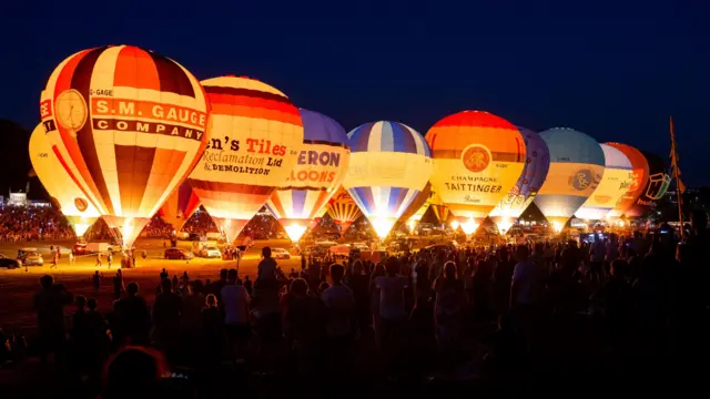 A large crowd is seen at night watching hot air balloons that are tethered on the ground fire their burners which are creating a glow