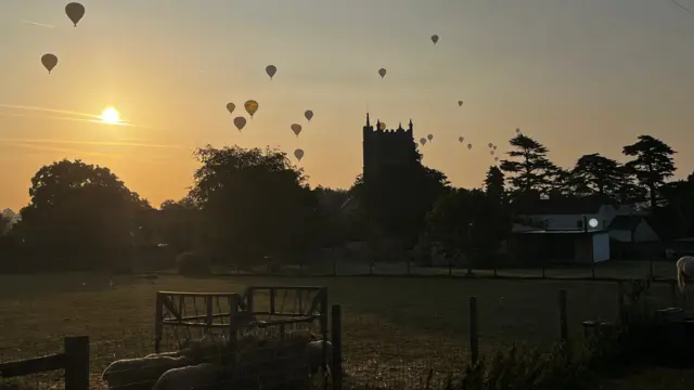 Silhouettes of balloons and the sun in the sky above a church and farmland