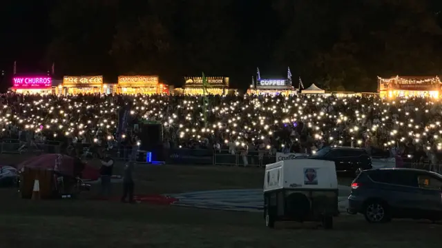 The crowd at the Bristol Balloon Fiesta hold their phones aloft in the darkness creating hundreds of pinpricks of light